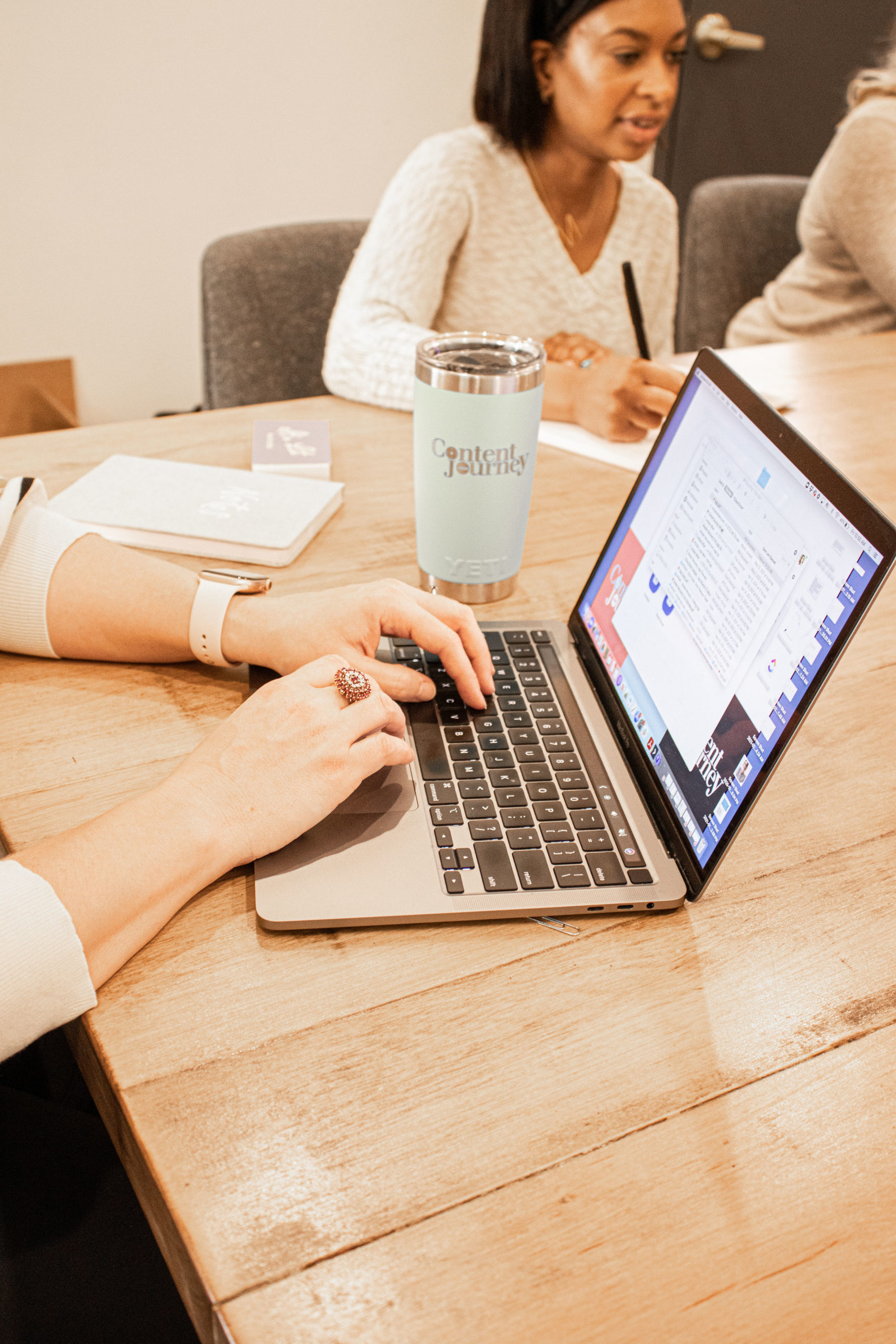 Hands typing on a laptop with a woman in the background