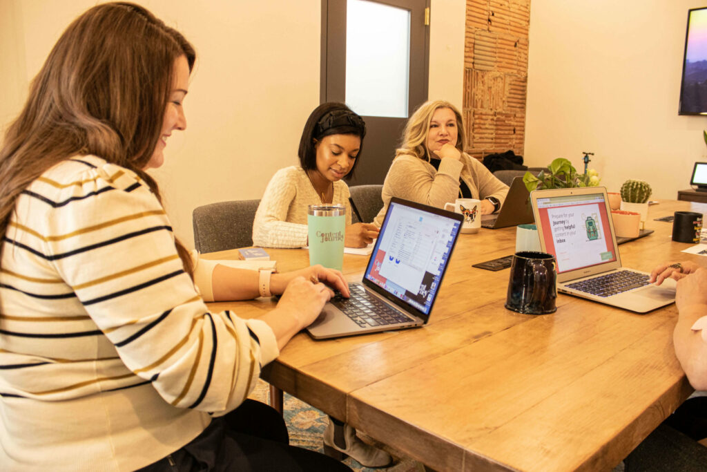Three women sit at a long table, computers and coffee mugs are on the table top.