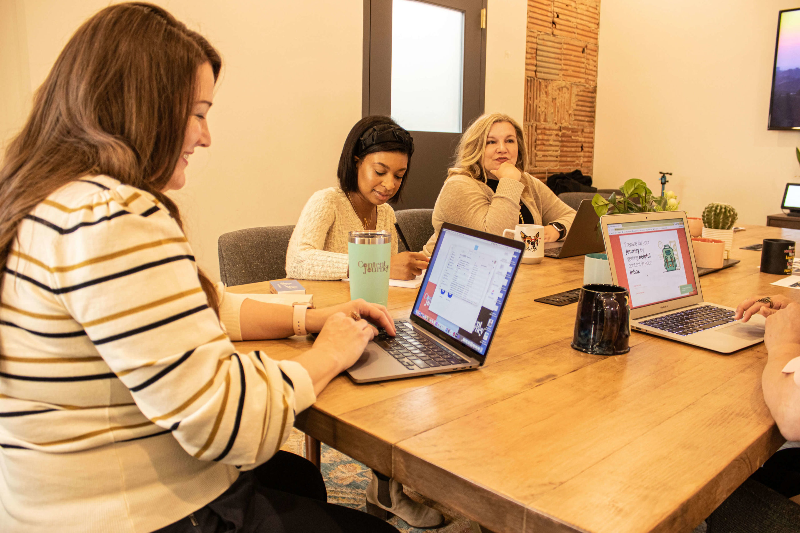 Three women sit at a long table, computers and coffee mugs are on the table top.