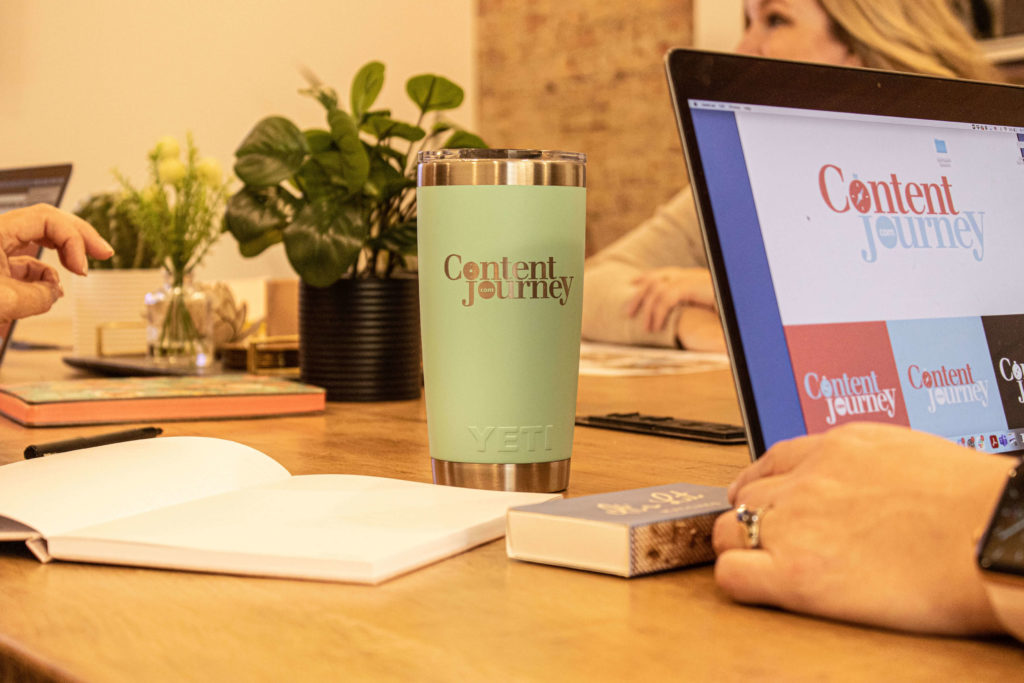 A computer and a coffee mug sit on a table where women are working, the logo for Content Journey is displayed