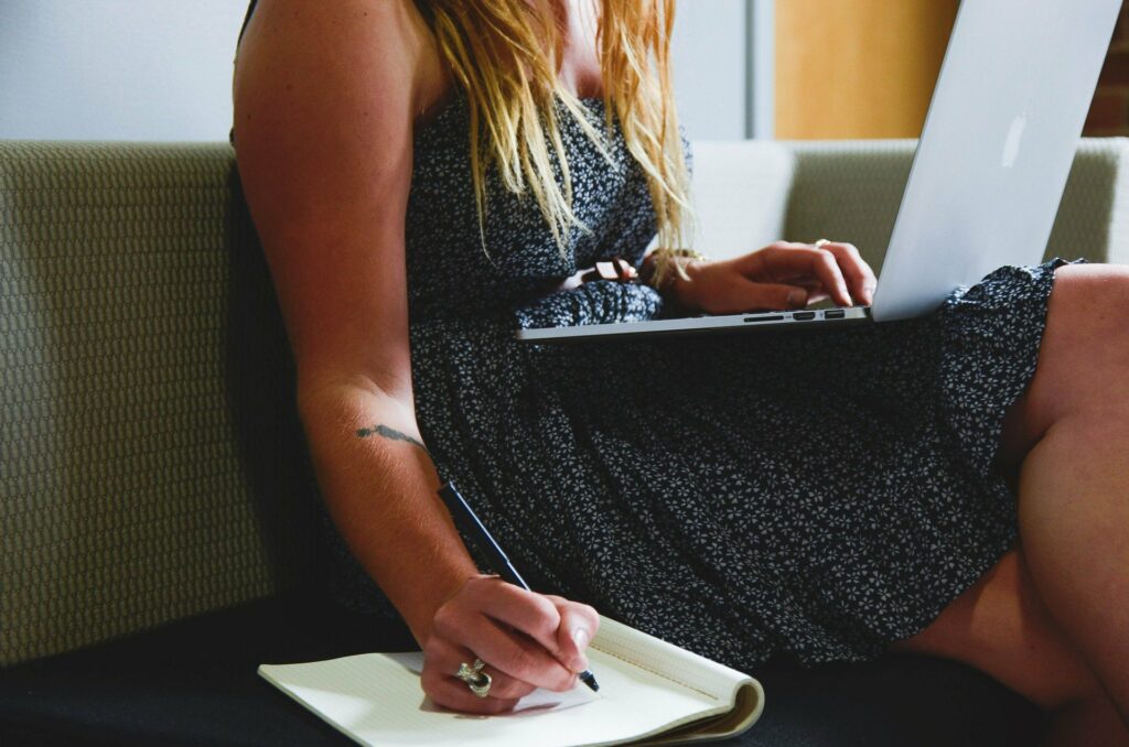 A woman writing in a notebook and typing on a computer at the same time.