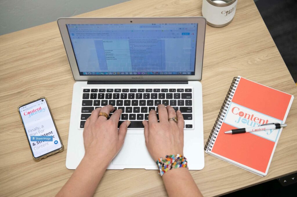 Hands typing on a laptop computer with a cell phone and notebook on either side of the laptop