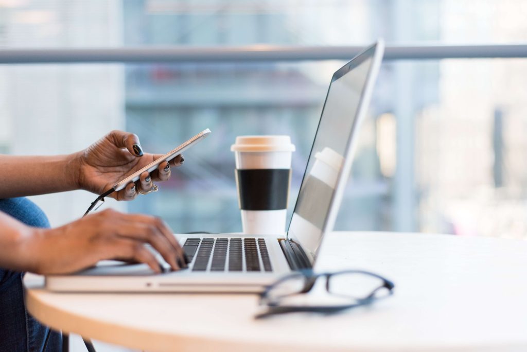 A woman's hands as she works on a laptop while holding an iPhone