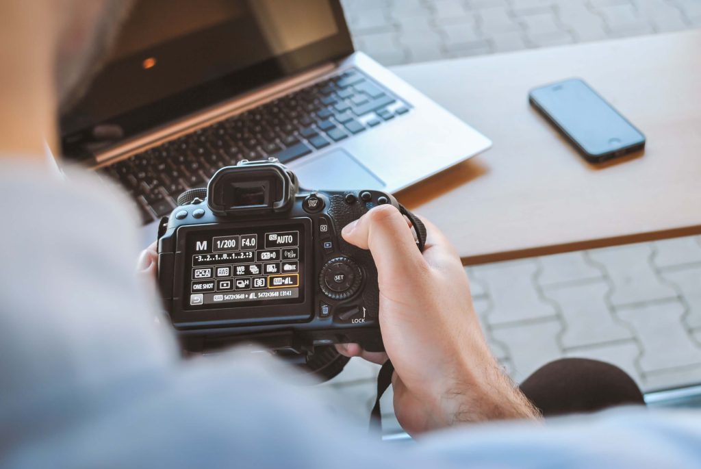 A close up of someone looking at digital camera settings with a laptop and iPhone in the background